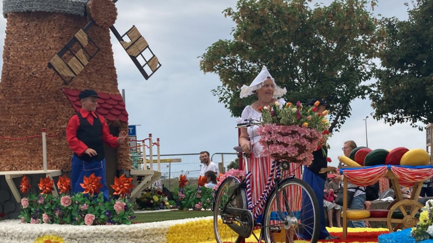 A woman stood on a float wearing a hat which is Dutch-themed. She has a bike with flowers and there are people at the front watching on.