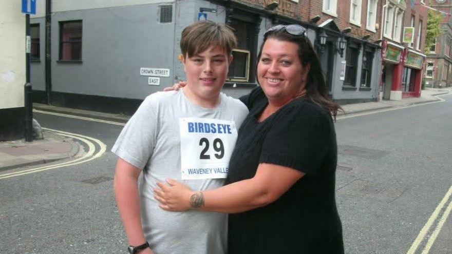 Connor, aged about 12, wears a grey T-shirt with a race number pinned to his chest. It says "Birds Eye 29 Waveney Valley". He is standing in a street with his mother, Toni. who is wearing a black top and had her arm round him. Both are smiling.