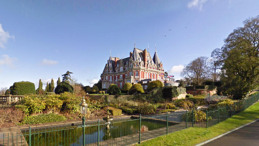 A 19th-century house built in the style of an elaborate French château. The property has large grounds with ponds and large hedges. The image has been taken on a sunny day. 