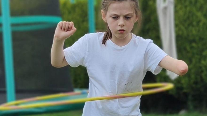 Zoey stands in a garden while making a hula hoop move around her waist. She has a grey T-shirt on and black shorts and is in front of a trampoline

