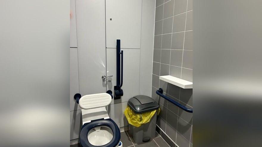 A toilet with a white backboard. A blue handrail is beside the toilet and a white shelf is attached to the wall next to it. A bin is in between the toilet and shelf.