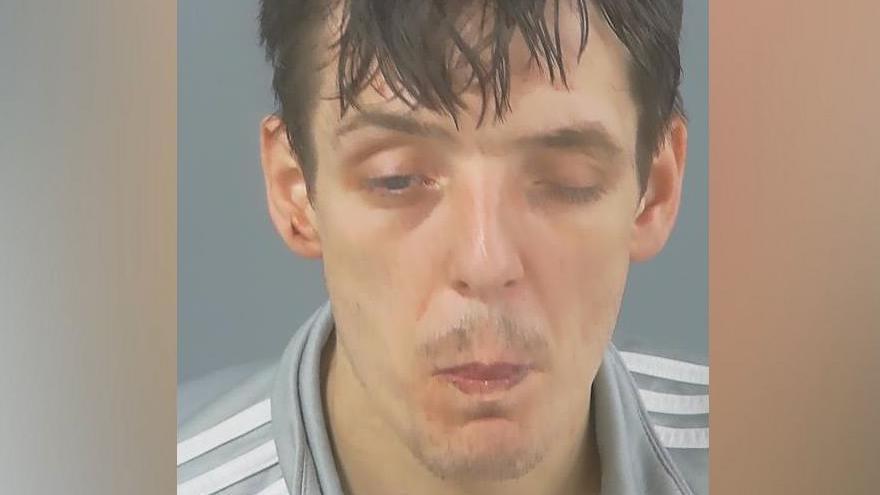 In a custody photo, Ashley Lear has one eye closed and is pursing his lips. He has messy, dark hair and a slight moustache and wears a light grey top with white stripes.