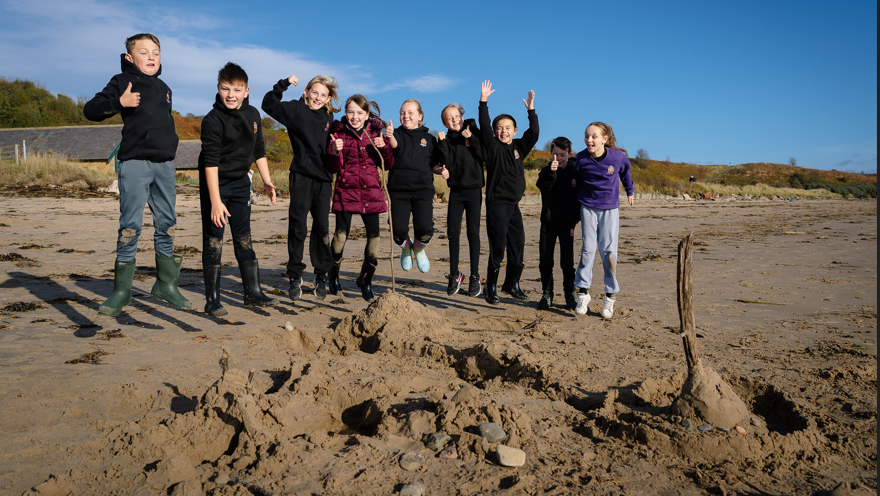 Eight children jumping in the air on a beach. They are standing in front of a piece of beach art they have created. It is a sunny day and two sheds are visible behind them. 