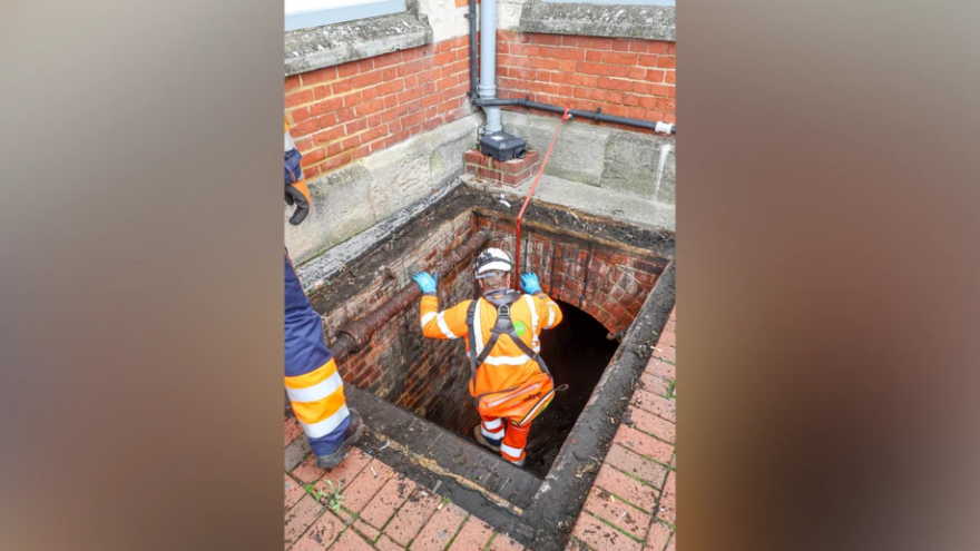 A large square bricked opening in the ground with a workman in a high-vis jacket being lowered in on a safety harness