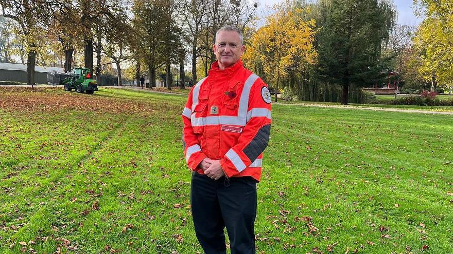 David Copeland from Norfolk Fire and Rescue Service stands surrounded by grass in the park. He is wearing an orange high-visibility jacket and black trousers. 