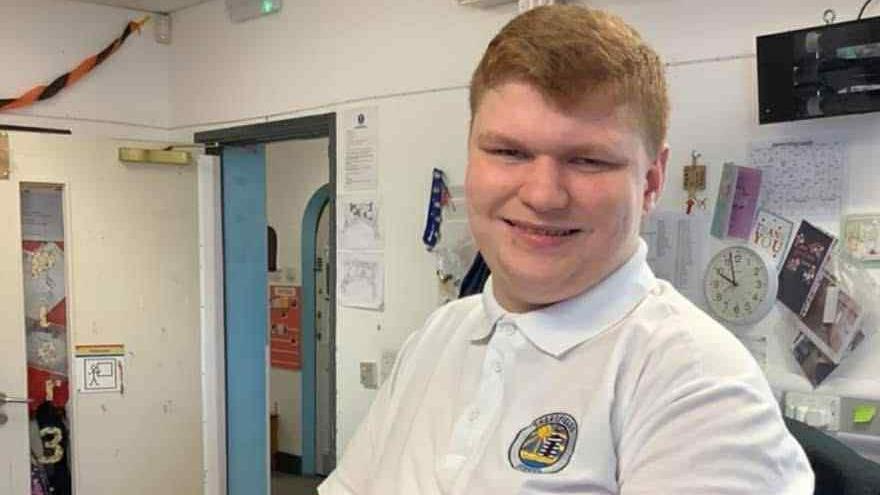 Ralph smiling inside a classroom with various notes and a clock pinned to a whiteboard. He is wearing a white polo shirt with the school's blue and yellow crest on it.