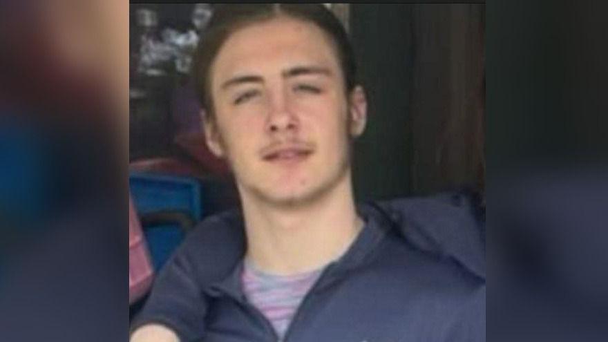 Kailem is smiling and looking at the camera. He has brown hair and is wearing a dark blue jacket and a pink and grey top.