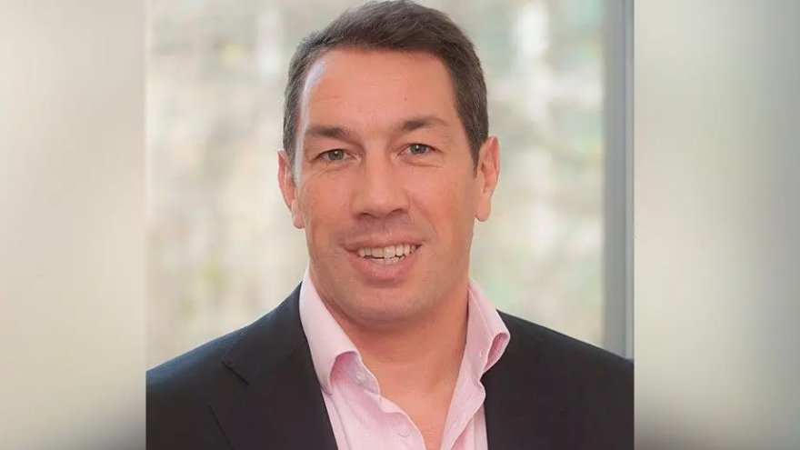 Tom Voyce in a formal photograph smiling at the camera and wearing a blue suit and pink, open-necked shirt.