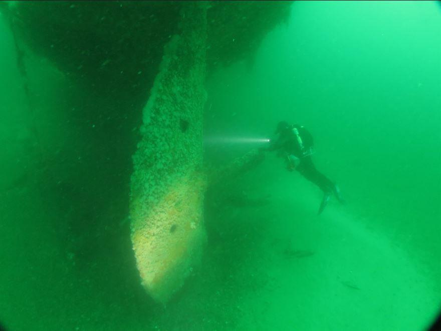 A man diving on the wreck of the Liverpool. The water is lot green and the ship's rudder can be seen in the fore of the scene.