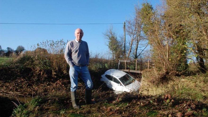 Dr Leslie Spiers is standing on the bank of the river, wearing wellie boots, jeans with a shirt and jumper. He has his hands in his pockets and is looking at the camera. The drivers white car is wedged beneath the bridge in the background.