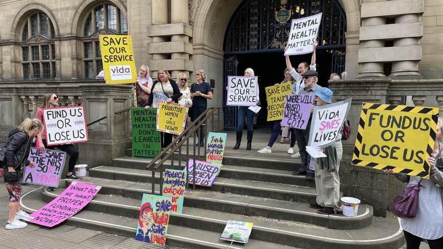 11 women and one man stand outside Sheffield Town Hall holding placards reading 'Fund us or lose us' and 'Vida saves lives'.  