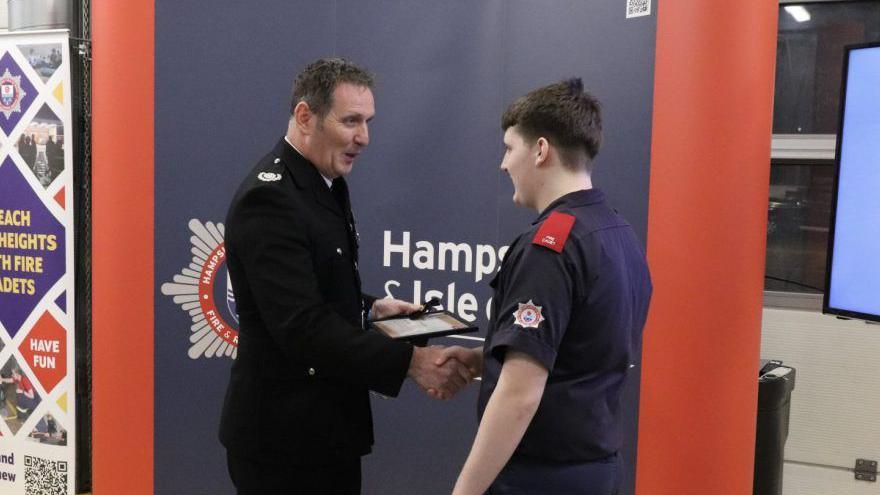 Robert in fire cadet uniform being handed a certificate and shaking the hand of a man in senior fire service clothing.