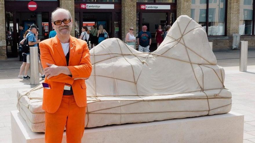 Gavin Turk, who is wearing an orange suit, standing in front of the Ariadne sculpture. The sculpture is made of grey stone and placed on top of a large rectangle block. The sculpture looks like a woman who has been covered with a sheet and rope. In the background is Cambridge railway station. Passengers are entering and leaving the station. The railway building is brick with lots of windows.