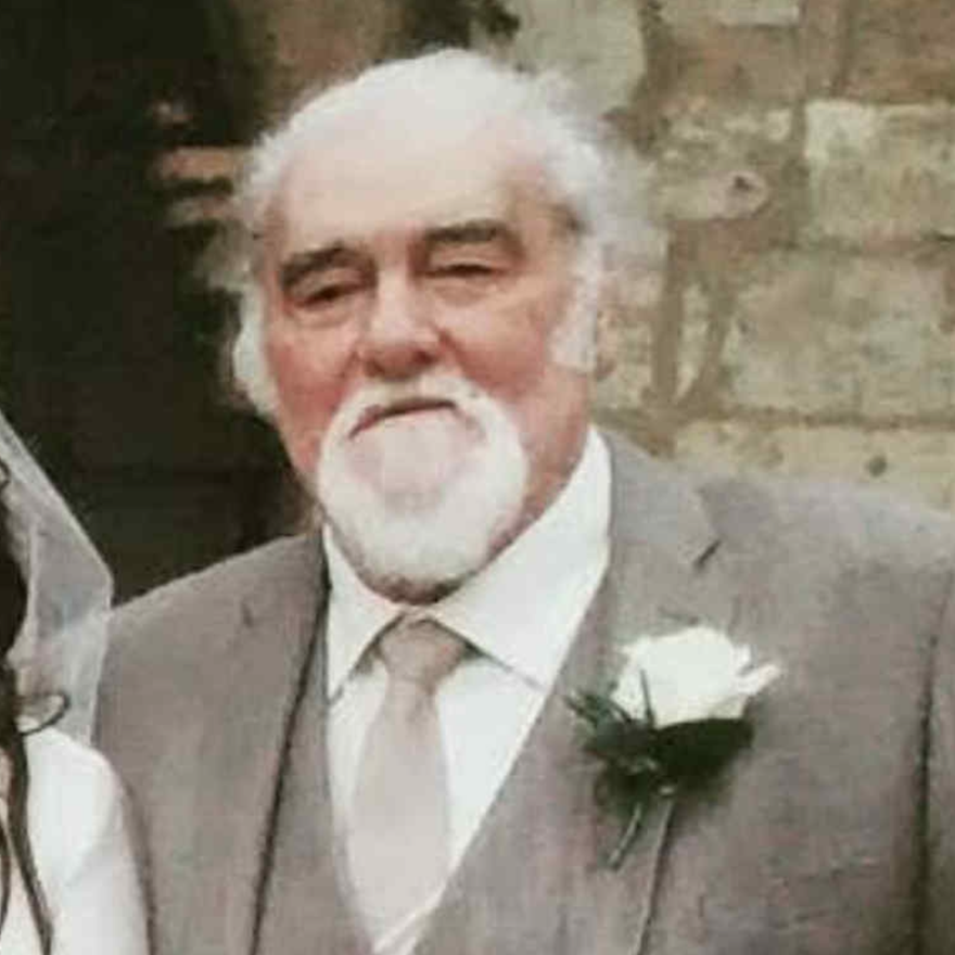 Robert has grey hair and a grey beard. He is wearing a grey suit and tie over a white shirt and there is a white flower pinned to his left lapel. He is standing in front of a church.