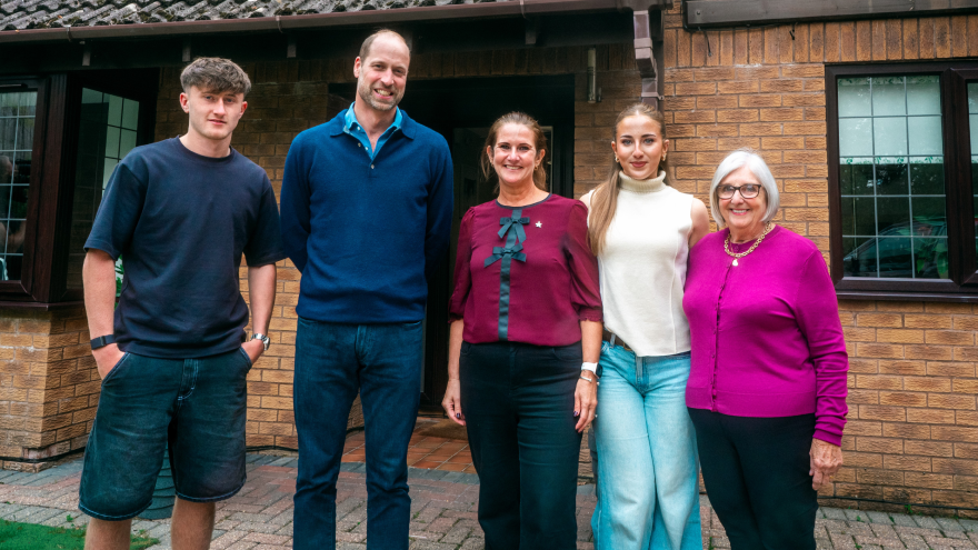 Prince William stood outside Rhian's front door with her two children, Holly and Isaac and her mother, Chris. Isaac, is on the left and wears a navy t-shirt with navy shorts and stands next to William. Rhian is next to William, with Holly stood next to her wearing a cream turtle neck jumper and blue jeans. Chris, on the far right, wears a pink knitted cardigan and black trousers. They are smiling looking at the camera.