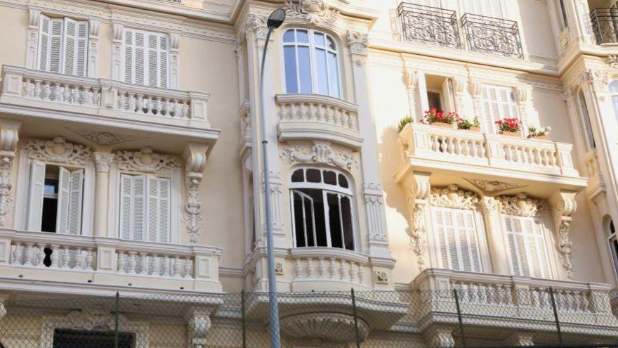 The damaged window of a light yellow residential building, which has balconies and ornate carvings.