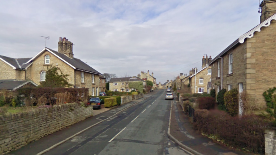 A Google streetview image of The Village, which is a road in Thorp Arch, Wetherby. The road goes uphill. A few cars are parked on the road in the distance, but most houses have driveways. The houses look large and stone-built. The plants and bushes around the houses are will maintained.