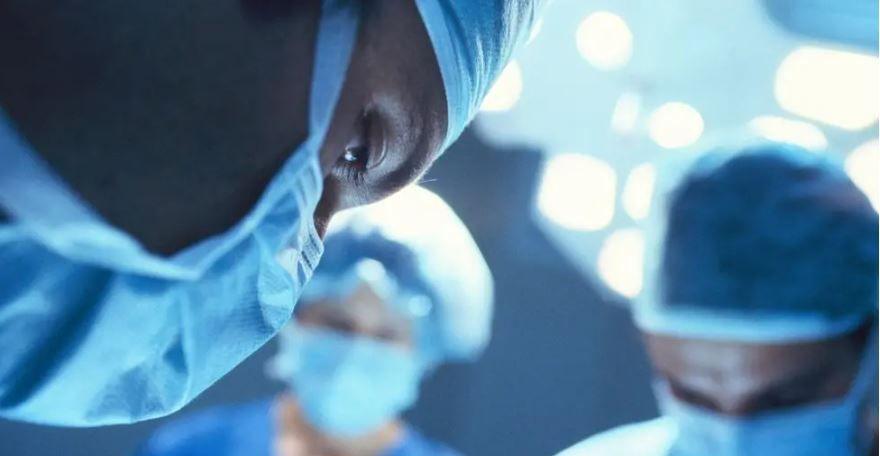 A surgeon wearing a mask and surgical hat in the foreground, with two other members of the surgical team wearing similar clothes in the background. All are looking down as they operate.