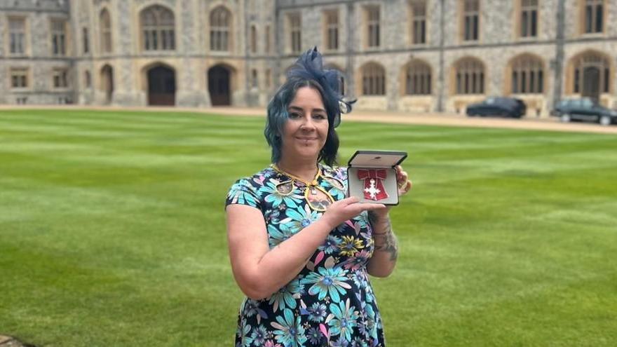 A woman with blue hair is wearing a dress with light blue flowers on it. She is smiling at the camera while holding up her MBE medal with its red ribbon.