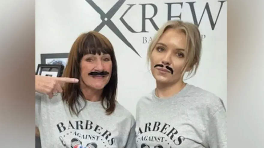 Two female barbers with fake moustaches wearing company uniform of a grey t-shirt with a poster for their shop, Krew Barbers, in the background