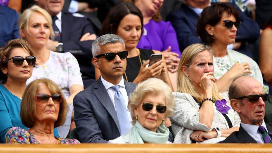 Sadiq Khan, wearing a blue suit and light blue tie and sunglasses, sits among a group of people at Wimbledon.