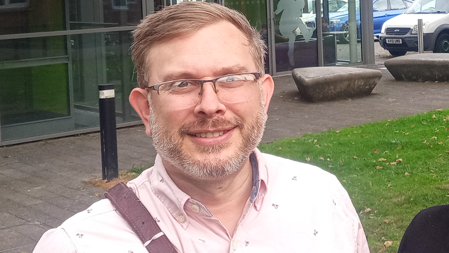 A man with blonde hair, glasses and a beard, wearing thin-framed glasses and a pale pink shirt standing in an area partly paved and partly covered in grass, in front of a glass-fronted building