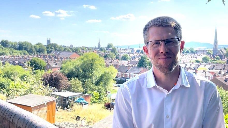 A man, who has short brown hair, a stubbly beard and is wearing glasses and a white shirt, is standing in front of a view of the city of Worcester with houses, steeples and greenery in the background.