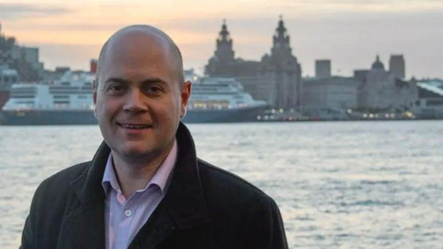 Liverpool council leader Liam Robinson standing on the waterfront in Wirral, with Liverpool's Three Graces visible across the Mersey behind him