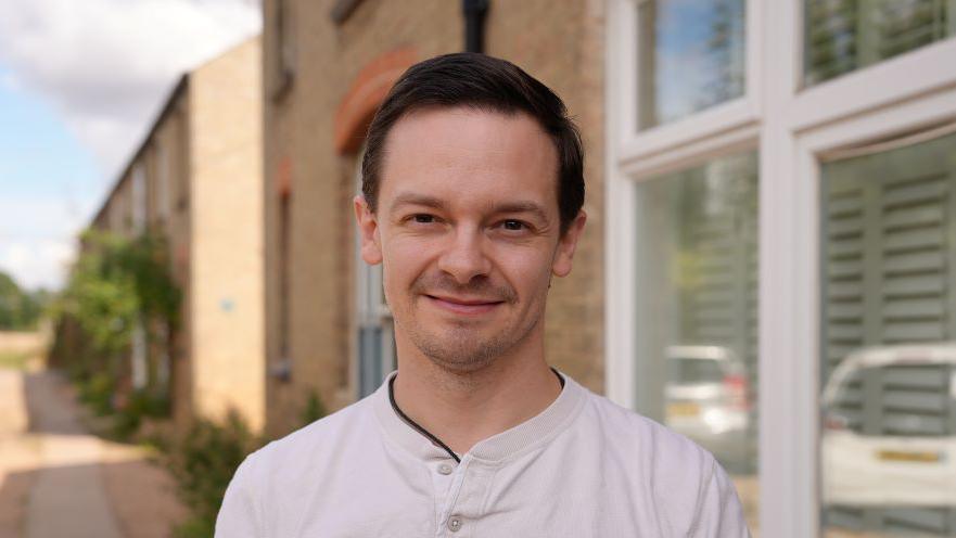 Chris Tuczemskyi, standing in a shaded part of a street. He is smiling and wearing a white, collarless shirt. 