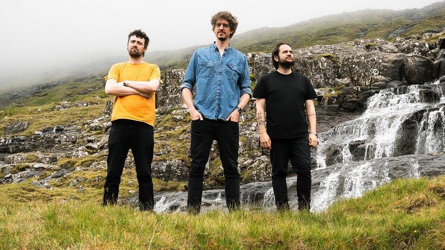 Three men stand on a mountain in front of a waterfall. The man on the right is wearing a yellow t-shirt and black jeans and is standing with his arms folded. The man in the middle is wearing a blue denim shirt and black jeans with his hands in the front pocket. The man on the right wears a black-t-shirt with black jeans and has his hands by his sides.