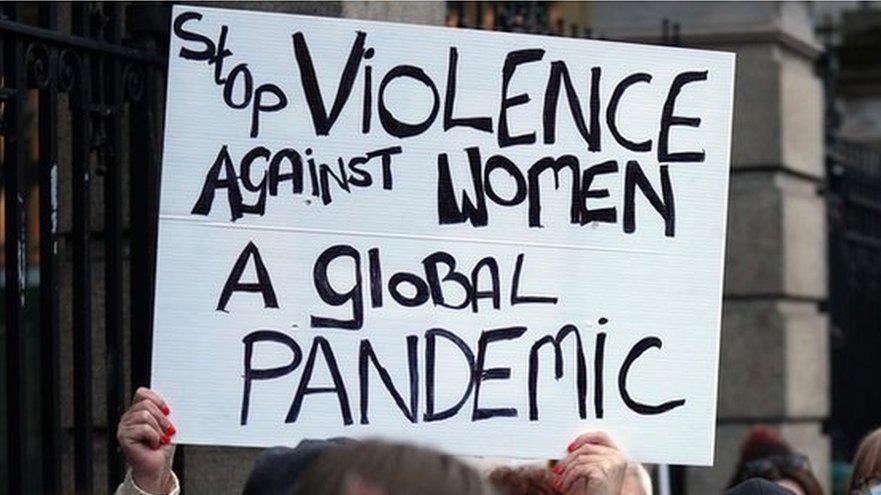 A woman holding up a homemade sign at a protest which says "Stop violence against women. A global pandemic."