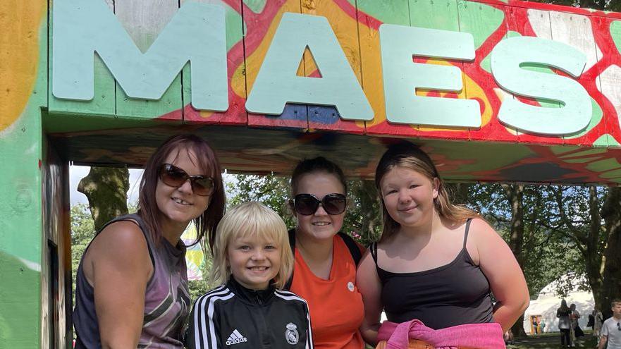 Rachel stood to the left of the photo. She is wearing sunglasses, short red hair and a purple top, stood next to eight-year-old Owen who has shoulder length blond hair and is wearing an Adidas black and white jumper, stood next to 20-year-old Morgann who is wearing her brown long hair in a ponytail and she is wearing sunglasses and then Izabel is stood next to her who has long blond hair and wearing a black tank top with a pink jumper tied around her waist. Trees can be seen in the back ground and one of the festival tents poking through the trees