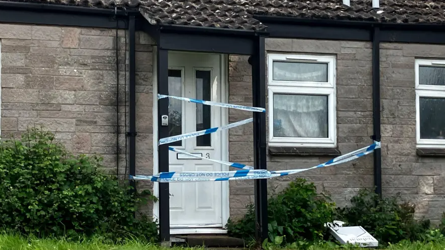 A white door to a bungalow in Glastonbury is covered with police tape.