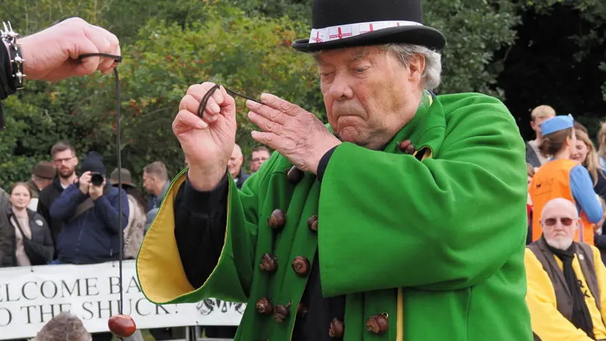 David Jakins takes aim at a conker being held on string. Mr Jakins has a look of concentration on his face. He is wearing a black hat and a green tunic.