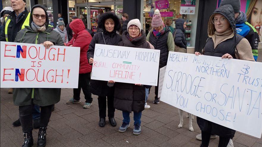 Three women holding placards standing on a street. One woman is wearing a brown coat and glasses and her placard says:  "Enough is Enough".
The woman in the middle is wearing a white woollen hat and a dark coat. Her placard reads:  Causeway hospital rural community lifeline".
The woman on the right is wearing a black and tan coat with the hood up and she has glasses. Her placard reads Scrooge or Santa, You choose".