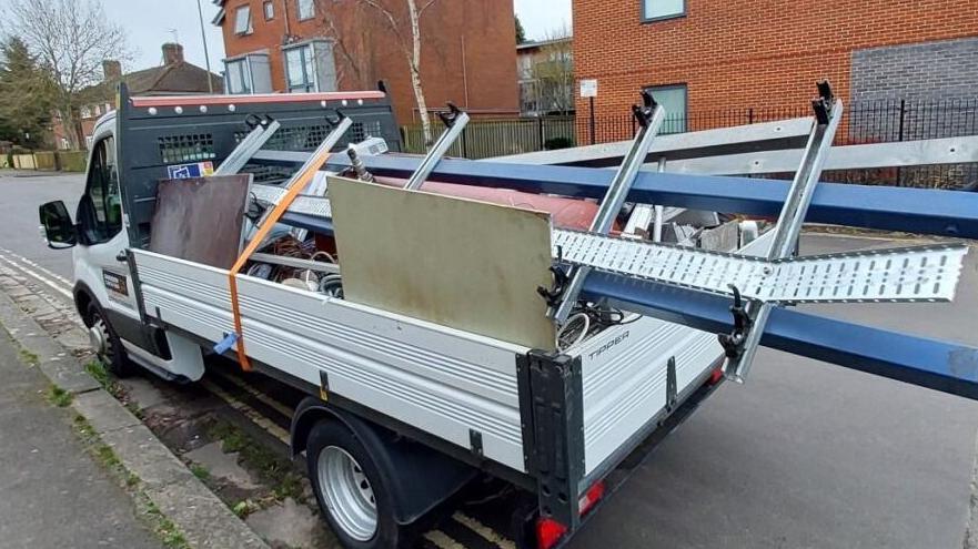 A small truck, parked on the side of a residential road, has metal bars protruding a long way out the back. The load of miscellaneous waste is tied down with a single, taut yellow strap.