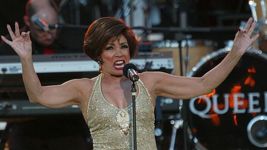 Shirley Bassey, wearing a gold sequinned dress with arms outstretched, performing on stage. 