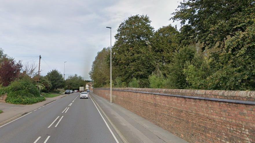 Road heading into Towcester with a path and patches of grass on the left and a brick wall with trees behind on the right. A white car is heading towards the camera.