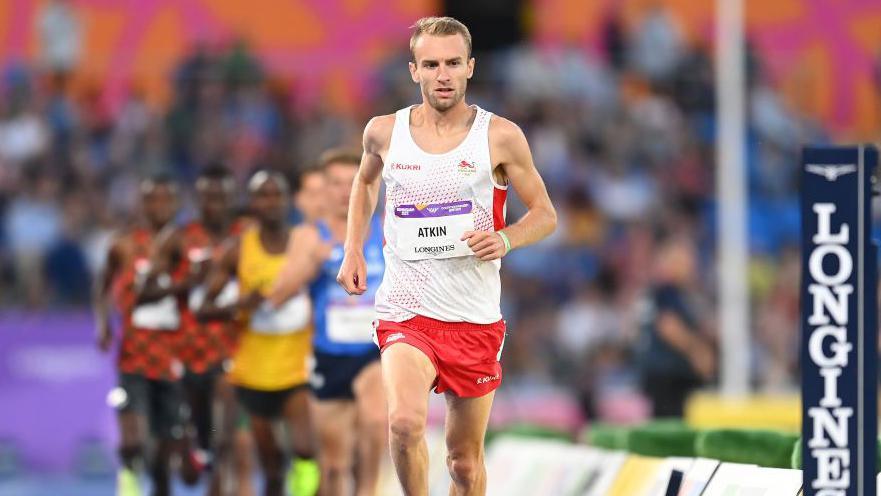 Sam Atkin, wearing a white vest and red shorts, runs at a track event in a stadium. He leads a field of competitors wearing vests of various colours.