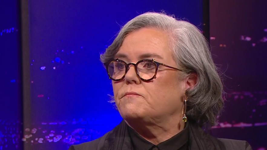 Rosie O'Donnell looks to her right while on the Late, Late Show. She has grey hair down to her shoulders and black-rimmed glasses and is wearing a dark jacket and shirt