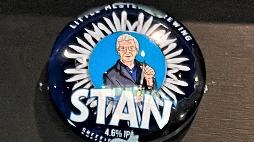 A circular blue badge depicting a cartoon image of a man with grey hair and glasses, Stan Shaw, holding a knife with a circle of assorted blades around him