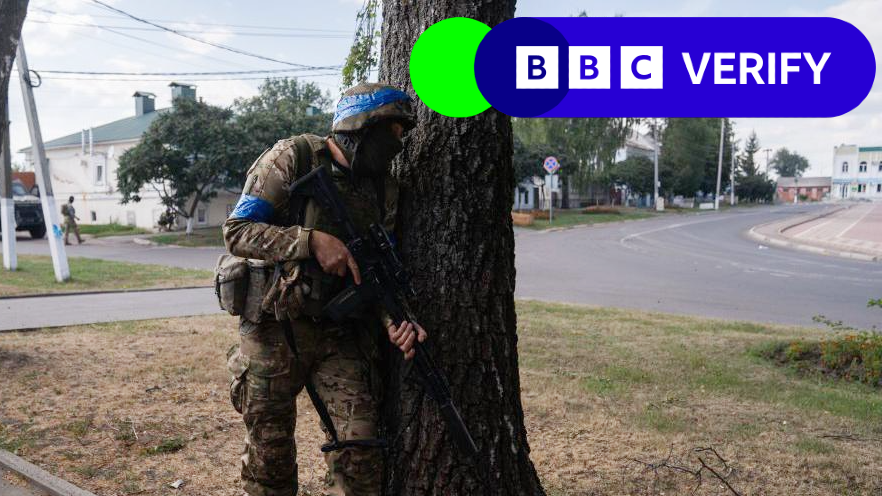 A Ukrainian soldier fighting in Russia's Kursk border region. He is carrying an assault rifle and is hiding behind a tree, while wearing a helmet and military fatigues. 