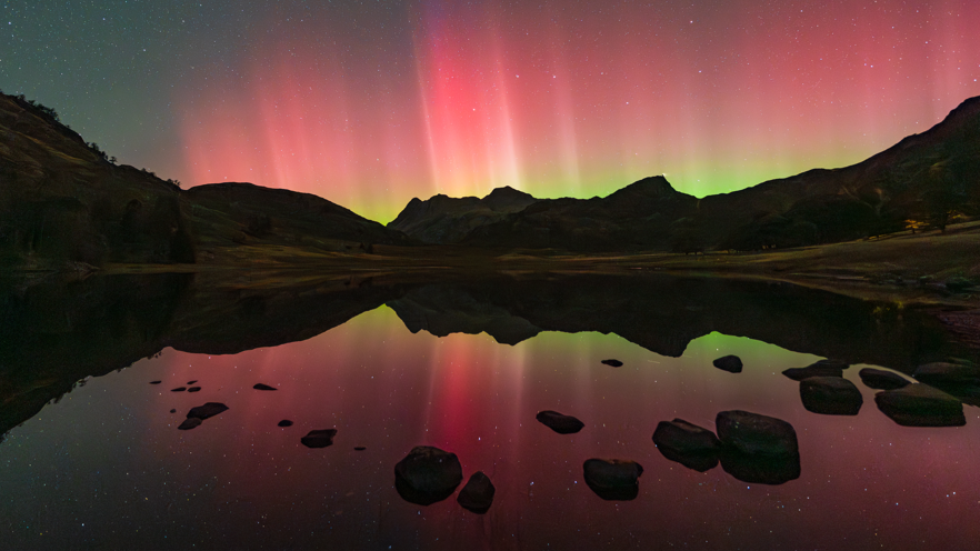 A lake in the foreground with pink and yellow light reflected in the water then the cliffs stretching away and the whole sky full of pink and yellow 