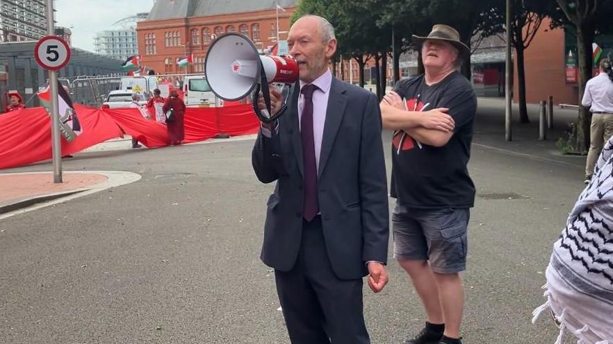 Newport East MS John Griffiths addressing protesters with a megaphone. The Megaphone is red and white. Protesters are holding a long piece of red cloth in the background, partially surrounding the Senedd.