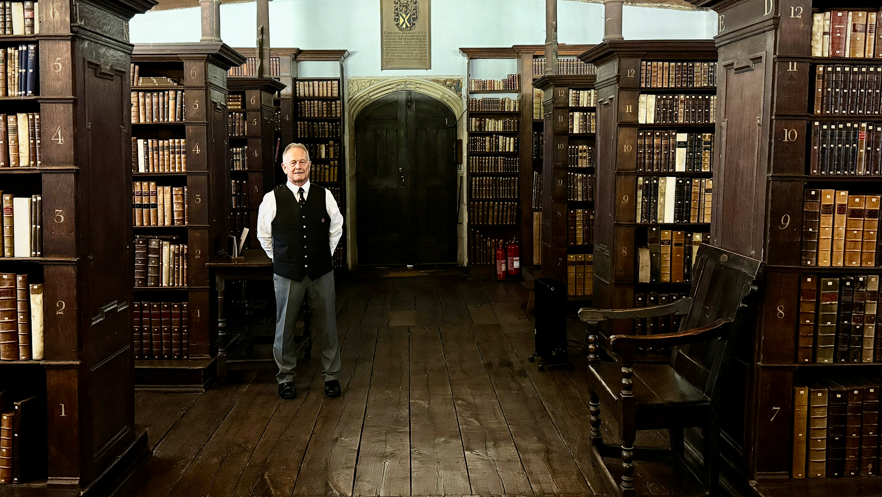 Dean Allen stood to the left of a wooden door which is set against a stone arch. Dean who has his hands behind his back and is standing to face the camera while smiling. He is in a dark library surrounded by books on large, dark wooden shelves. The floor is also dark and wooden, matching the bookcases and a bench to the right of the image.
Dean wears a Porter's outfit which consists of a black bowler hat, white shirt, black tie, black waistcoat and grey suit trousers. The black tie and breast pocket of the waist coat have the Jesus College logo. 