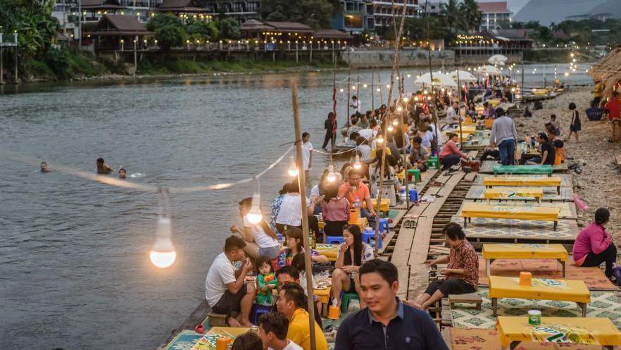 People drink and enjoy food on a riverbank in the town of Vang Vieng in Laos. There as some people swiming in the river and a string of lights highlights dozens of people sat on tables.