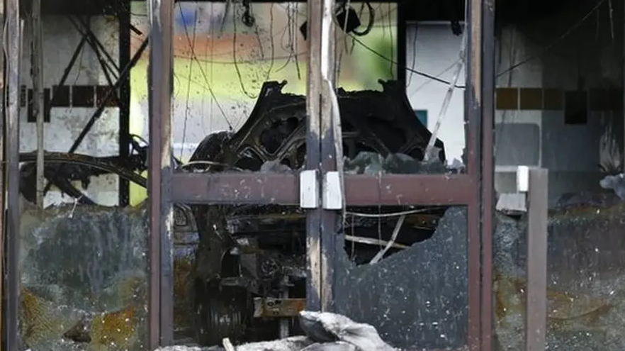 A clearly fire-damaged HQ building in Crowmarsh Gifford after the fire in January 2015. The shell of the building remains badly burned and most of the glass has smashed or shattered. In the centre of the picture is a pair of doors, with metal door handles. They have no glass inside them, aside from a charred black pane in the bottom right of the two. The other side of the building is left open, so you can see grass and/or a hedge, but it is blurred. It is unclear if that is fire damaged or if a door has been opened