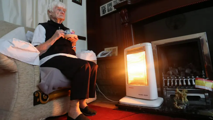 An elderly lady sitting on a sofa in a lounge holding a cup of tea in front of an electric fire. She is wearing a black gilet over a white top. She has black trousers and black shoes on. The lady has short white hair.