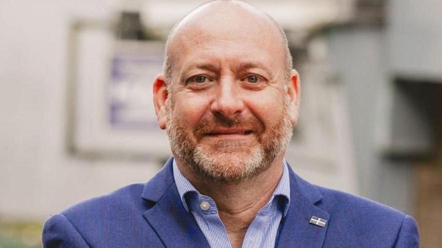 Labour MP for Camborne and Redruth Perran Moon stands wearing a dark blue blazer and a light blue shirt with a Cornish flag badge on his lapel. He has a grey and giner beard and short hair.