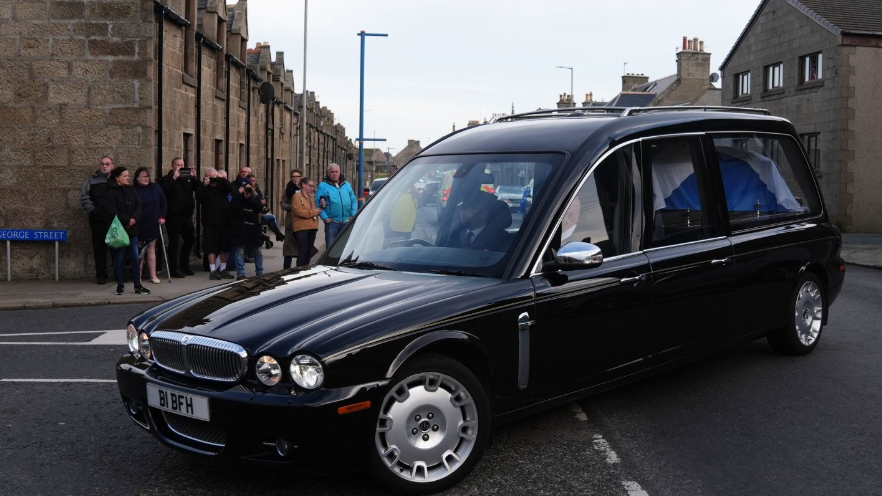 Black hearse turns corner of street with small crowd watching from pavement in front of historic row of houses.